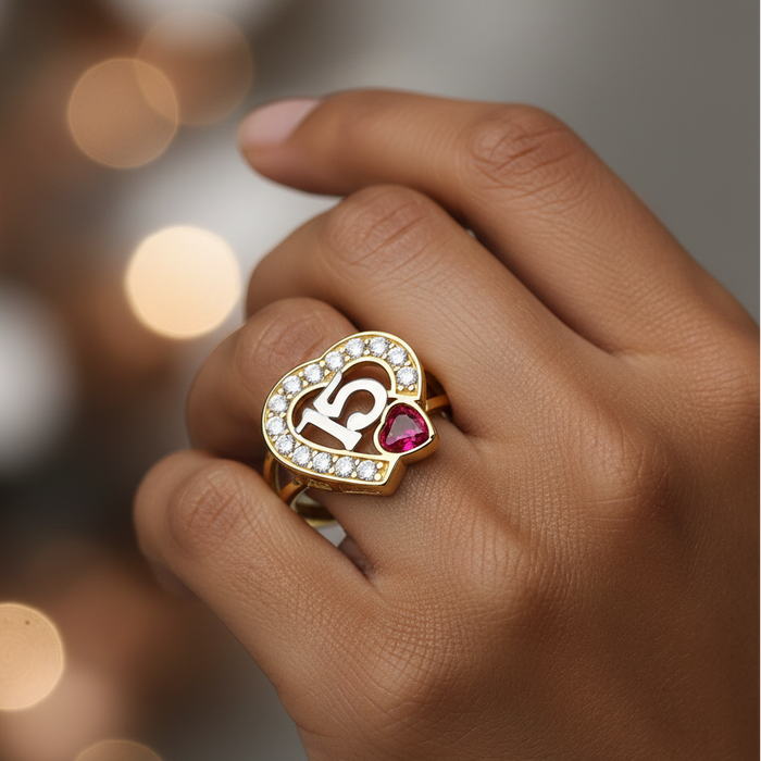 Hand wearing a gold ring with a heart design and red gemstone against a blurred background
