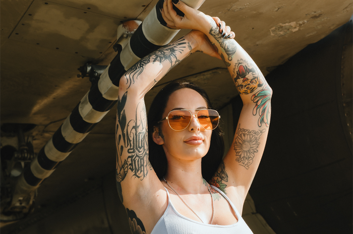 Woman with tattoos wearing Aviator sunglasses with a polished gold frame and amber lenses under an F-14 airplane.