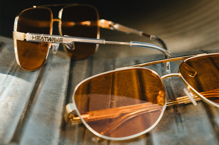 Gold-framed sunglasses with a polished gold frame and amber lenses, featuring “Heatwave” branding, displayed on a reflective surface.