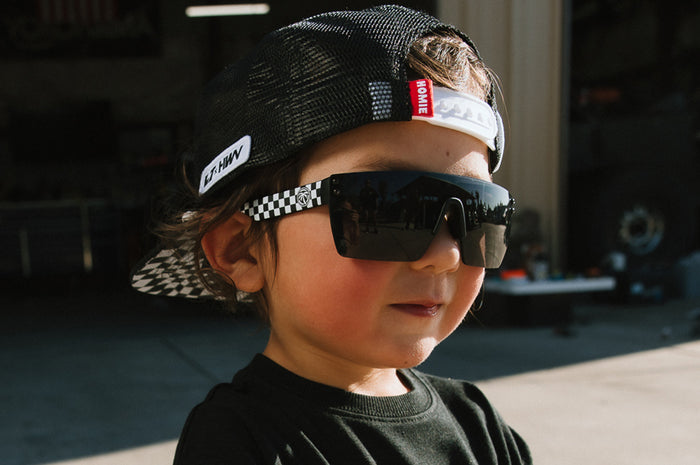 Young boy wearing the Kids Lazer Face Sunglasses: Little Trouble X Heat Wave Checkered Flag with black lens outside.