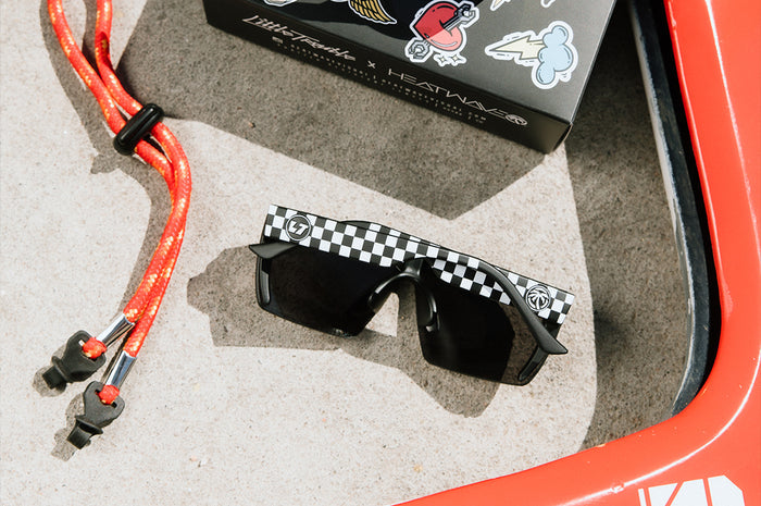 Backside shot of the the Kids Lazer Face Sunglasses: Little Trouble X Heat Wave Checkered Flag on a concrete floor next to the strap and product box.