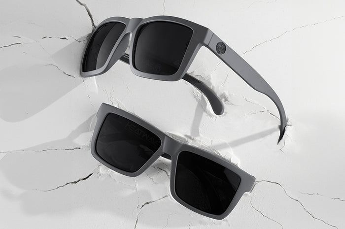 2 pairs of Heat Wave Visual Vise Sunglasses with satin grey frame and black lenses on cracked white surface.